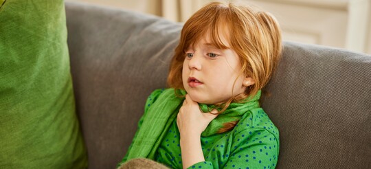 Mädchen mit Halsschmerzen – GeloRevoice® zur Linderung von Halsschmerzen bei Kindern. Ein kleines Mädchen mit roten Haaren sitzt auf einem Sofa und hält sich den Hals, während es einen Schal trägt. Sie wirkt erschöpft und hat offensichtlich Halsschmerzen. Eine Decke liegt über ihren Beinen, was auf Ruhe und Erholung hinweist.