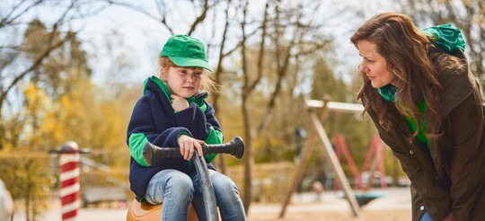 Kind mit Heiserkeit im Park – Tipps zur Linderung von Halsbeschwerden bei Kindern. Kind sitzt auf einem Spielgerät im Park und hält sich den Hals, während eine Frau sich zu ihm herunterbeugt und besorgt schaut.