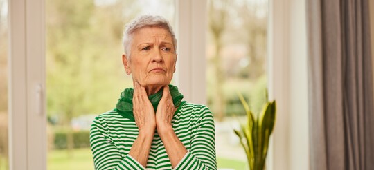 Ältere Frau mit Halsbeschwerden – mögliche Anzeichen einer Mandelentzündung und Tipps zur Linderung Ältere Frau in grün-weiß gestreiftem Oberteil steht vor einem Fenster und fasst sich mit schmerzverzerrtem Gesichtsausdruck an den Hals, was auf Beschwerden wie Halsschmerzen oder eine Mandelentzündung hindeutet.