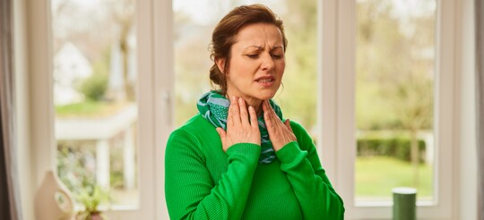Frau mit Halsbeschwerden – mögliche Linderung mit GeloRevoice® Halstabletten Frau im grünen Pullover hält sich mit schmerzvollem Gesichtsausdruck den Hals und leidet offensichtlich unter Halsbeschwerden.