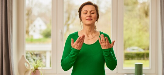 Entspannung und Stimmtraining für eine starke Stimme – Übungen für die Stimmpflege Frau in grünem Oberteil steht mit geschlossenen Augen und hebt die Hände vor ihrem Gesicht, während sie eine Atem- und Stimmübung durchführt. Im Hintergrund ist ein Fenster mit Gartenblick zu sehen.