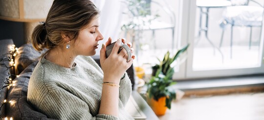 Schwangere Frau mit Halsschmerzen sitzt auf der Couch und trinkt Tee: In der Schwangerschaft können bestimmte Hausmittel die Linderung unterstützen.