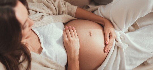 Schwangere Frau mit Halsschmerzen legt Hände auf ihren Bauch: Sie ist sich unsicher, was sie in der Schwangerschaft anwenden kann.