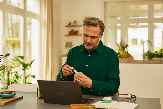 Ein Mann sitzt am Tisch mit einem Laptop und hält eine Packung GeloRevoice® Halstabletten in der Hand, während er aufmerksam die Verpackung betrachtet. Im Hintergrund eine moderne Küche.