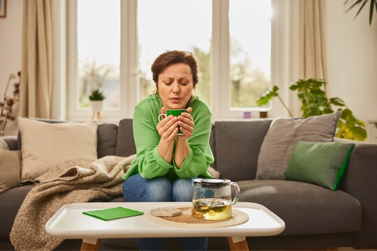 Eine Frau sitzt auf einem Sofa und hält eine dampfende Tasse Tee in beiden Händen. Vor ihr steht eine Teekanne auf einem Tisch. Sie entspannt und ruht sich aus.