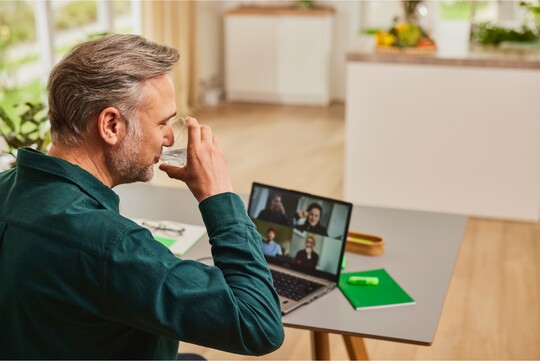 Ein Mann trinkt ein Glas Wasser, während er an einer Videokonferenz auf seinem Laptop teilnimmt. Er sitzt in einem hellen Raum mit Pflanzen und Notizbuch vor sich. Die Szene betont die Bedeutung der Flüssigkeitszufuhr zur Linderung von Halsschmerzen oder einer Rachenentzündung.