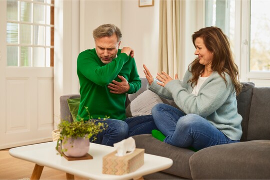 Ein Mann hustet in seinen Arm, während eine Frau auf Abstand geht und versucht, sich vor den Tröpfchen zu schützen. Beide sitzen auf einem Sofa in einem gemütlichen Wohnzimmer. Auf dem Tisch stehen eine Pflanze und eine Taschentücherbox.