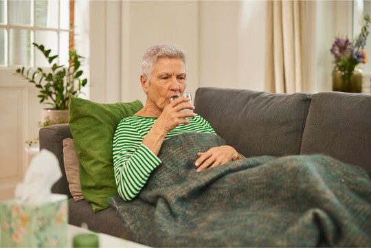 Ältere Frau sitzt in eine Decke eingewickelt auf einem Sofa und trinkt ein Glas Wasser, um Mundtrockenheit zu lindern. Neben ihr steht eine Taschentuchbox und eine Kerze.