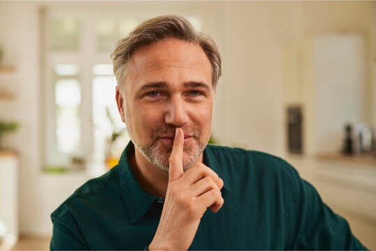 Ein Mann mittleren Alters mit grauem Haar und Bart lächelt leicht und hält einen Finger an seine Lippen, als Zeichen für Ruhe und Stille. Er trägt ein dunkelgrünes Hemd und befindet sich in einem hellen Raum. Die Geste deutet auf die Bedeutung von Stimmschonung als Teil der Stimmhygiene hin.