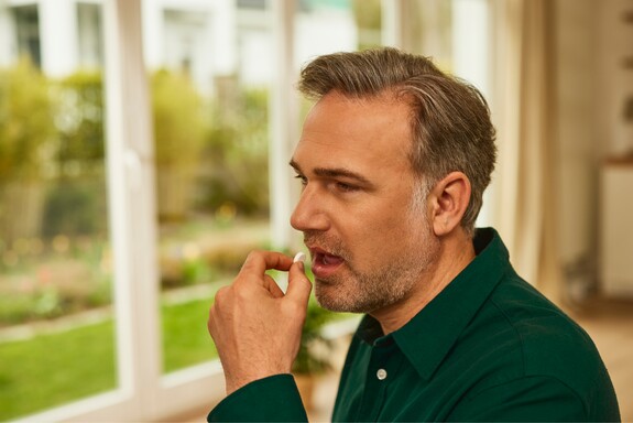 Mann mittleren Alters nimmt eine GeloRevoice® Halstablette ein, im Hintergrund große Fenster mit Blick in den Garten.