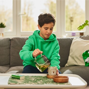 Ein wärmender Kräutertee und GeloRevoice® – sanfte Linderung für Halsbeschwerden Junge in grünem Hoodie gießt Kräutertee aus einer Glaskanne in eine Tasse auf einem Wohnzimmertisch. Neben ihm liegt eine Packung GeloRevoice® Halstabletten, die für Unterstützung bei Halskratzen und Heiserkeit sorgt.