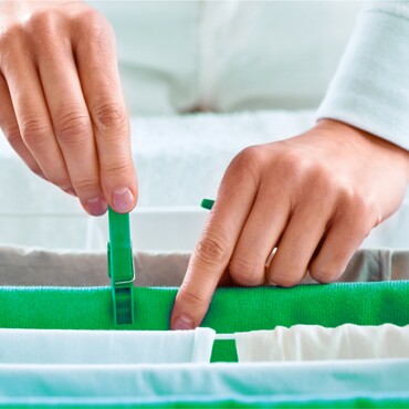 Wäsche aufhängen für frische Luftfeuchtigkeit – ein einfacher Schritt für ein angenehmeres Raumklima Nahaufnahme von Händen, die Wäsche auf einer Leine mit grünen Wäscheklammern befestigen. Der Fokus liegt auf den Fingern und der Klammer, während die frisch gewaschene Kleidung aufgehängt wird.