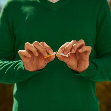 Eine Person in einem grünen Pullover bricht eine Zigarette in zwei Hälften. Die Szene symbolisiert das Aufhören mit dem Rauchen, um die Gesundheit des Halses zu fördern und Mandelentzündungen sowie Halsschmerzen zu vermeiden.