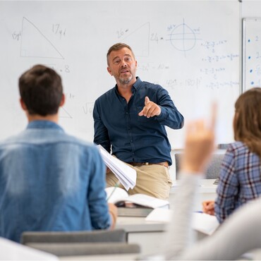 Lehrer im Klassenzimmer erklärt eine mathematische Aufgabe und zeigt auf einen Schüler. Im Hintergrund sind mathematische Formeln und Zeichnungen an der Tafel zu sehen. Der tägliche Einsatz der Stimme im Unterricht erfordert Schutz und Pflege, wie ihn GeloRevoice® bietet, um Heiserkeit und Stimmprobleme zu vermeiden.