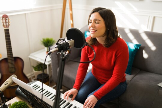 Schutz der Stimme beim Singen – Wie GeloRevoice® Sängerinnen bei Stimmproblemen unterstützt Junge Frau in rotem Pullover singt mit Leidenschaft ins Mikrofon in einem Heimstudio, begleitet von einem Keyboard. Im Hintergrund stehen eine Gitarre und ein Sofa, die Umgebung wirkt gemütlich und kreativ.