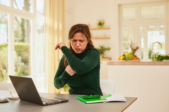 Symptome von Heiserkeit: Frau hustet am Schreibtisch und zeigt Anzeichen von Unwohlsein Frau in dunkelgrünem Pullover hustet in die Armbeuge und schaut angestrengt, während sie am Schreibtisch sitzt.