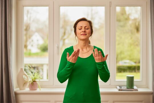 Entspannung und Stimmtraining für eine starke Stimme – Übungen für die Stimmpflege Frau in grünem Oberteil steht mit geschlossenen Augen und hebt die Hände vor ihrem Gesicht, während sie eine Atem- und Stimmübung durchführt. Im Hintergrund ist ein Fenster mit Gartenblick zu sehen.