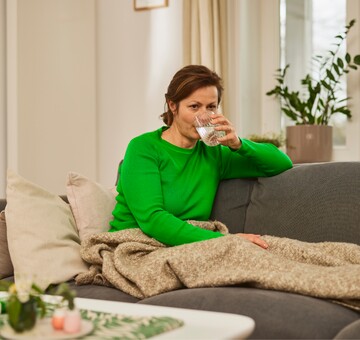 Frau trinkt Wasser auf dem Sofa – Flüssigkeitszufuhr als Teil der Stimmhygiene. Frau in grünem Pullover sitzt auf einem Sofa und trinkt ein Glas Wasser. Sie ist in eine beige Decke gehüllt, und im Hintergrund stehen Pflanzen, was eine ruhige und entspannte Atmosphäre schafft. Die Szene deutet auf Stimmhygiene und die Wichtigkeit der Flüssigkeitszufuhr für eine gesunde Stimme hin.