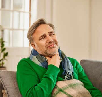 Schmerzen im Hals und Heiserkeit – Ursachen und Linderung bei Aphonie Mann mittleren Alters in grünem Pullover sitzt auf einem Sofa, hält sich den schmerzenden Hals und wirkt gequält. Er trägt einen Schal und scheint unter Heiserkeit oder Stimmverlust zu leiden.
