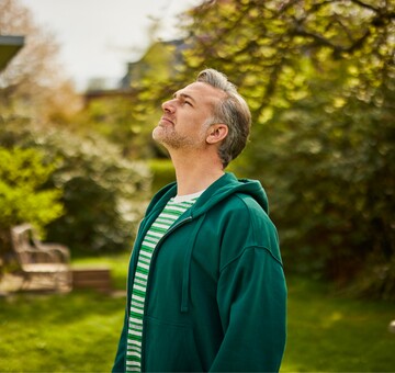 Entspannung und frische Luft im Garten – tiefes Durchatmen für mehr Wohlbefinden Mann mittleren Alters im grünen Hoodie steht draußen im Garten und blickt entspannt nach oben, während er tief durchatmet und die Natur genießt.