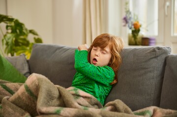 Hustendes Mädchen auf Sofa – GeloRevoice® zur Linderung von Reizhusten bei Kindern. Ein rothaariges Mädchen hustet in ihren Ellenbogen, während es auf einem Sofa sitzt, in eine Decke gehüllt.